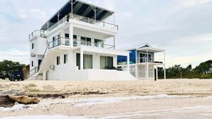 Une maison moderne à deux étages, située en bord de mer, dotée d’un balcon, de grandes fenêtres et d’un espace extérieur couvert.