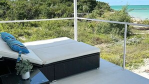 Un balcon doté d’un plafond blanc, d’un canapé en osier noir, d’une petite table avec une plante, et offrant une vue sur la plage et l’océan.