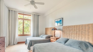 Een slaapkamer met twee bedden, een plafondventilator, een raam met gordijnen, een nachtkastje met een lamp, en een lijst met een afbeelding aan de muur.