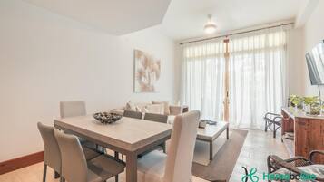 A modern living room with a dining area, featuring a wooden dining table, beige chairs, a sofa, and a large window with sheer curtains.
