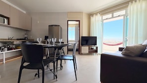 A modern living room with a sofa, a dining area with chairs and a table, a TV, and a view of the beach. - Tranquilidade e muito ventilado  (Aquiraz)