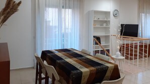 Un comedor con una mesa cubierta por un mantel estampado, rodeada de sillas. Se observa una estantería con diversos objetos y un televisor sobre un soporte.