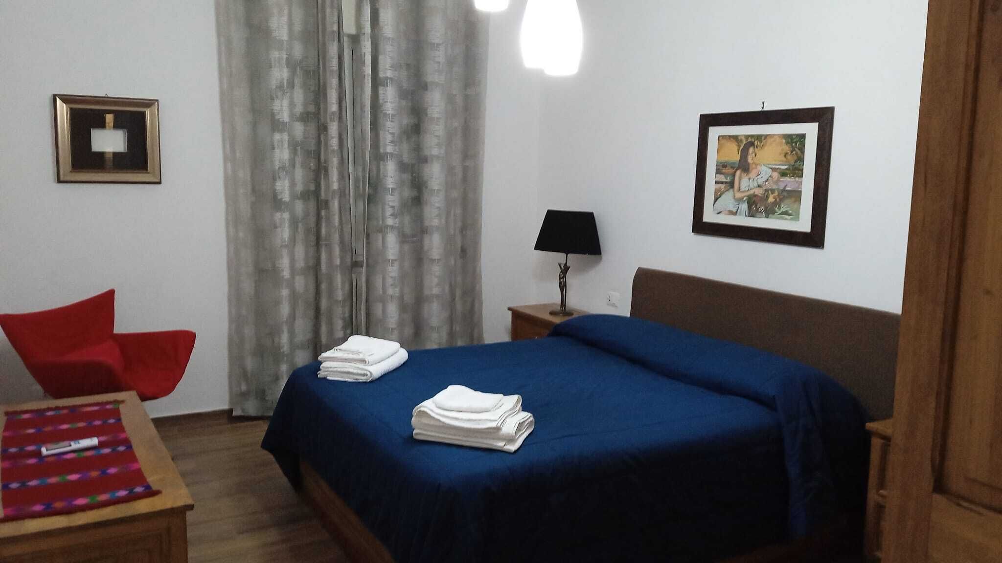A hotel room with a bed, a chair, a table, a lamp, and a picture on the wall.