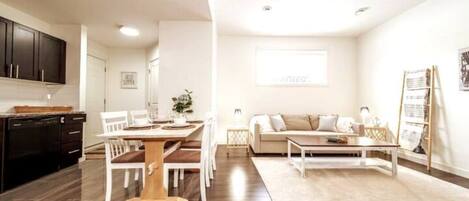 Une cuisine moderne avec des meubles de couleur sombre, un coin repas avec une table en bois et des chaises blanches, un salon avec un canapé et une table basse, et une grande fenêtre qui laisse entrer la lumière naturelle.
