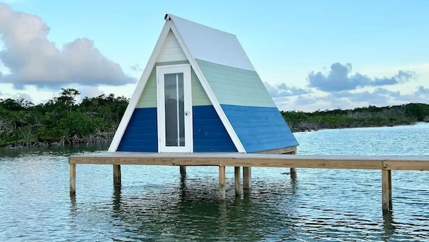 A small, triangular, elevated wooden structure with a glass door, situated on stilts over water, surrounded by vegetation.
