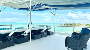 A covered outdoor seating area with lounge chairs and a wicker chair, overlooking a beach and ocean.