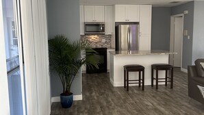 A modern kitchen with white cabinets, a stainless steel refrigerator, and a microwave. There is a potted plant and two bar stools.