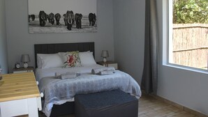 Un dormitorio con una cama, dos mesitas de noche, una lámpara de noche, una ventana que da a una valla y un cuadro colgado en la pared con elefantes.