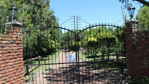Una entrada protegida con muro de ladrillo y portón metálico, que da paso a un sendero rodeado de follaje.
