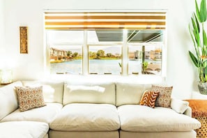 Sala de estar com sofá, almofadas estampadas, janela com persianas e vista para uma piscina e prédios.