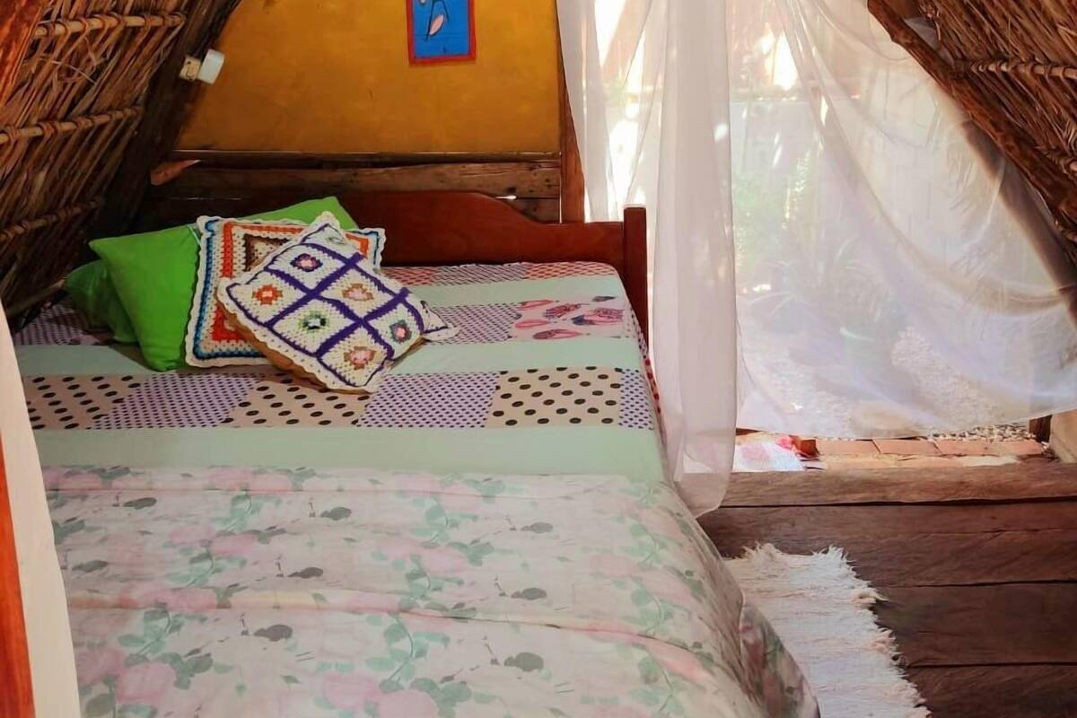 A thatched-roof room with a bed, a hanging blue and red decoration, and a white curtain.