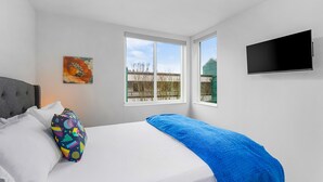 A bedroom with a bed, a colorful pillow, a blue blanket, a painting, and a TV on the wall. - Time Travelers Cowboys Sweet Suite (Seattle)