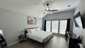 A bedroom with a bed, a desk, a chair, a ceiling fan, and large windows.