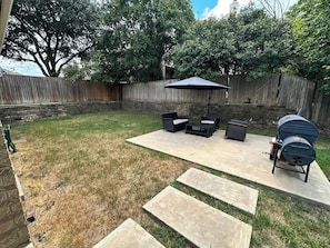 Ein Garten mit einer Beton-Terrasse, Gartenmöbeln, einem Grill und einem Holzzaun.