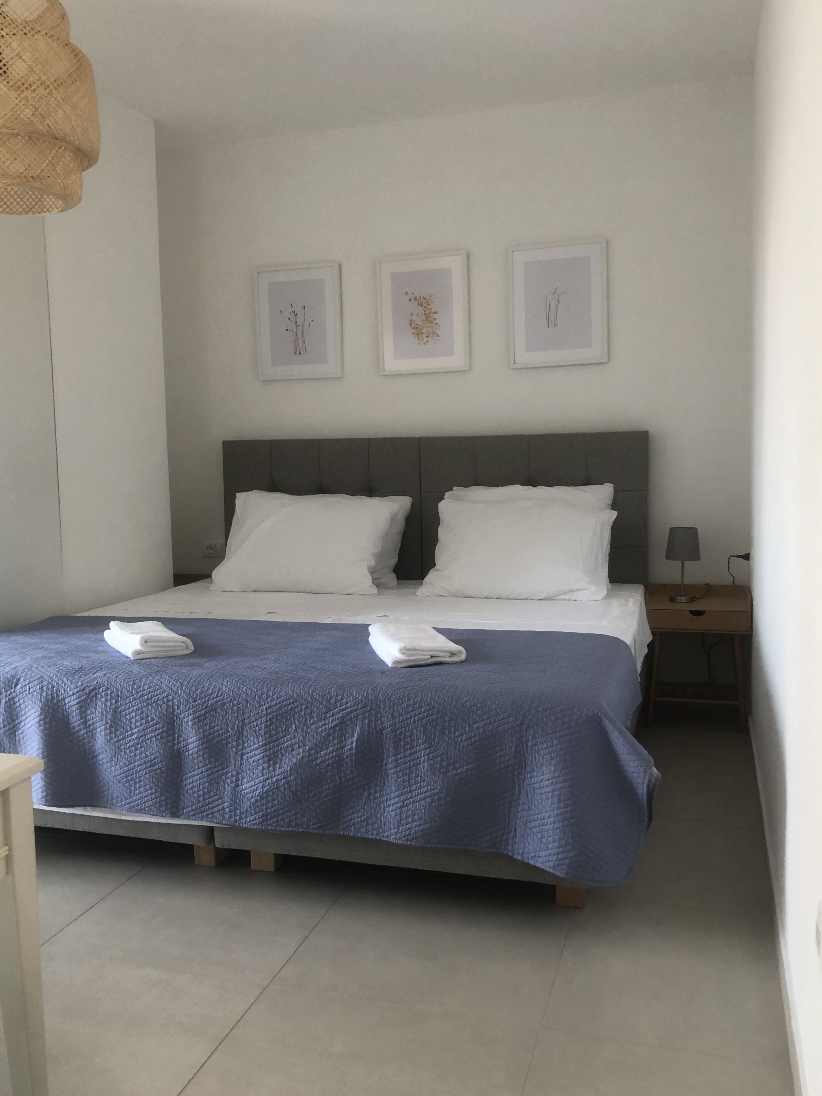 Ein ordentlich bezogenes Bett mit blauer Decke und weißen Kissen, ein Nachttisch mit Lampe und drei gerahmte Bilder an der Wand.