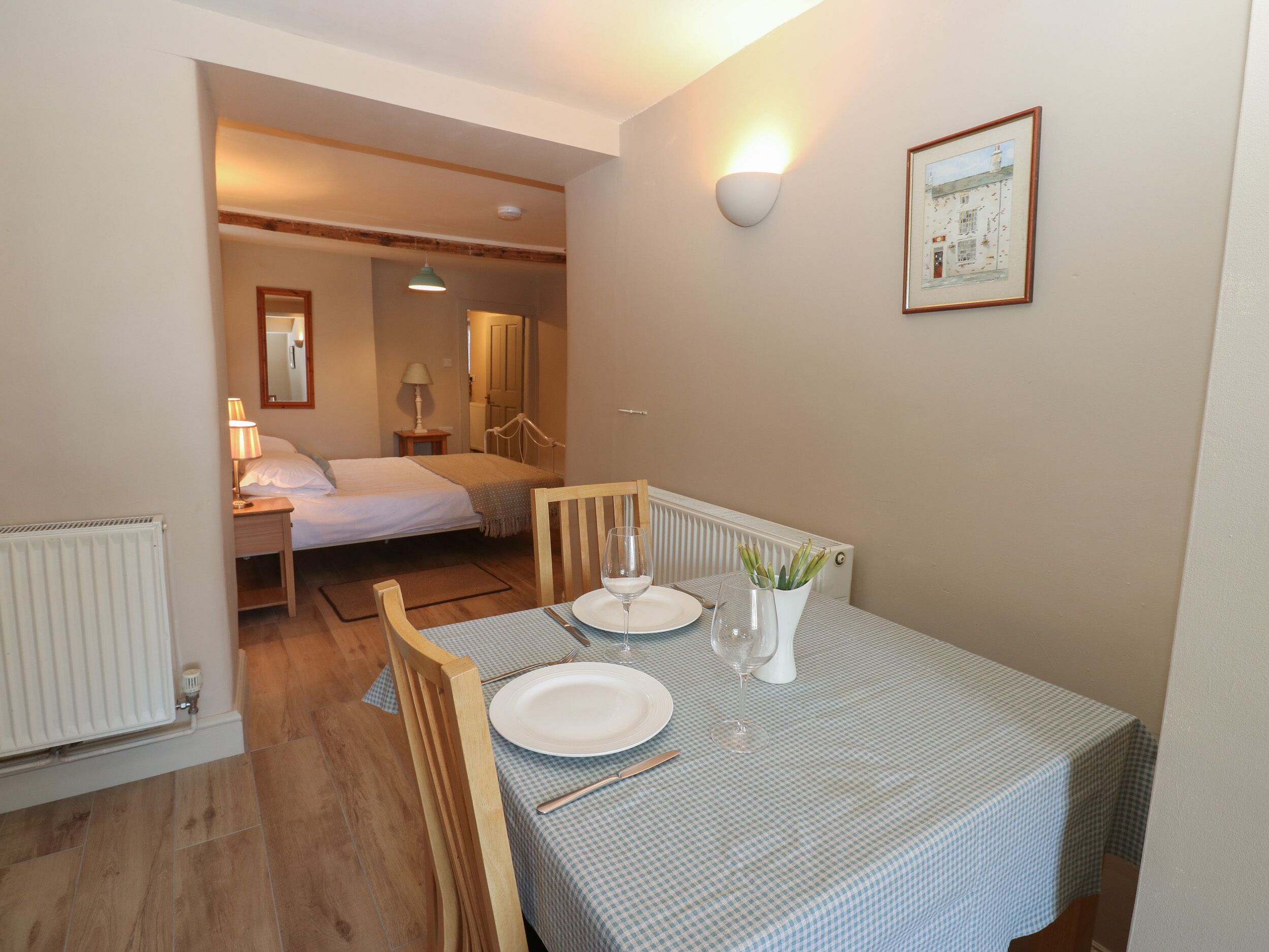Una habitación con una mesa de comedor dispuesta para dos personas, una cama con cabecera de madera, una mesita con lámpara y una lámpara de pared.