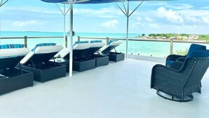 A covered outdoor seating area with lounge chairs and a wicker chair, overlooking a beach and ocean. - Beachfront 1 Bedroom/Pool/Kitchen/Breakfast/550SF (San Pedro)