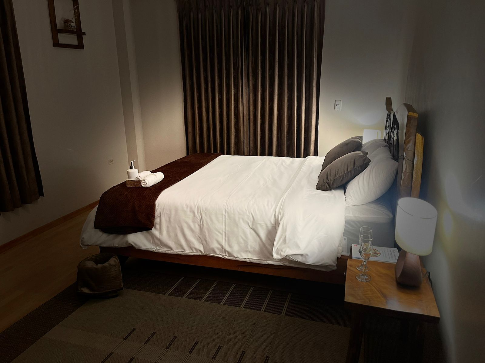 A neatly made bed with a white duvet and brown throw, a bedside table with a lamp, and a small rug on the floor.