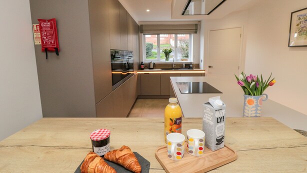 Une cuisine moderne avec une table en bois, deux croissants, une bouteille de jus, deux tasses à pois, un emballage de lait d’avoine et une couverture anti-feu accrochée au mur.