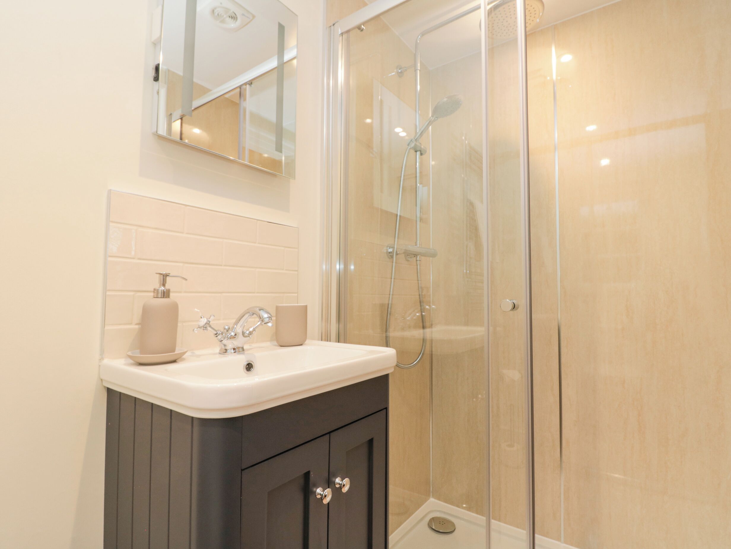 A modern bathroom with a glass-enclosed shower, a white sink with a soap dispenser, and a mirror above the sink.