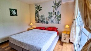 Un dormitorio con cama, dos mesitas de noche con lámparas, un gran mural con motivos tropicales en la pared y una ventana con cortinas.