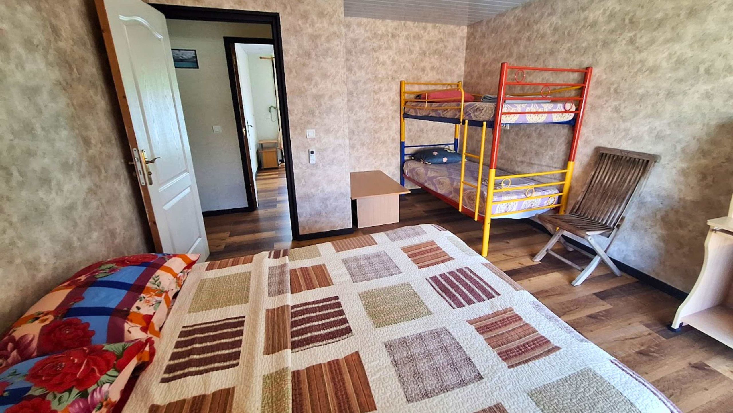 A room with bunk beds, a single bed with a floral comforter, a wooden chair, and a small table.