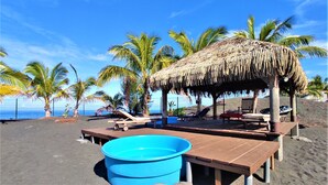 Una playa con una cabaña de techo de paja, bancos de madera y una piscina azul.