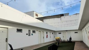 Un patio con césped artificial, mobiliario exterior, luces colgantes y un edificio con varias puertas.