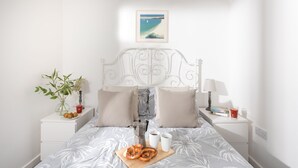 A neatly made bed with a grey floral bedspread, a wooden tray with pretzels and a cup, a framed picture on the wall, and a bedside table with a lamp and a glass of water. - Wheal Amelia Lodge (Helston)