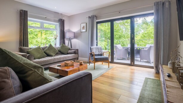 A spacious living room with a sofa, a coffee table, and a chair. There is a large window with curtains, a door leading to a balcony, and a lamp. - Bridge Howe (Ambleside)