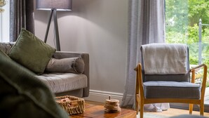 A living room with a grey sofa, a wooden chair, a floor lamp, and a window with curtains. - Bridge Howe (Ambleside)