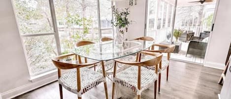 Un comedor con una mesa de vidrio rodeada de sillas de mimbre, ventanas amplias y un ventilador de techo.