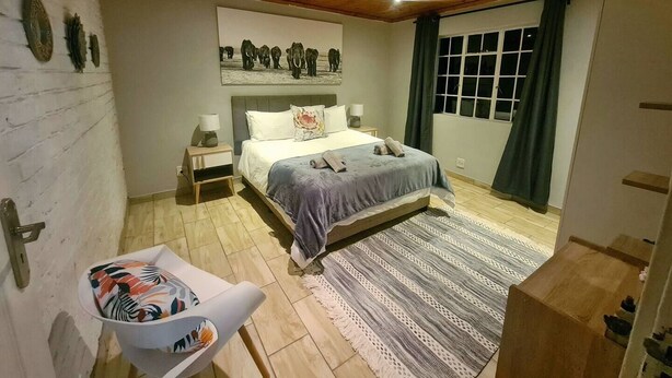 Een slaapkamer met een houten plafond, een bed met een deken van grijs en blauw, een nachtkastje met een lamp, een raam met gordijnen, en een wanddecoratie van olifanten.