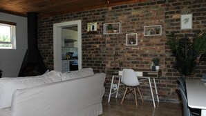 Een woonkamer met een witte bank, een eethoek met een witte stoel, een bakstenen muur, en een keuken die door een open deur zichtbaar is.