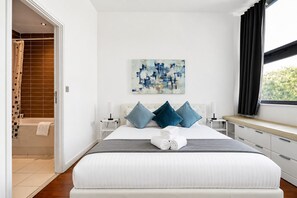 Une chambre moderne avec un grand lit, une fenêtre avec des rideaux, un tableau accroché au mur et une salle de bain visible à travers une porte ouverte.