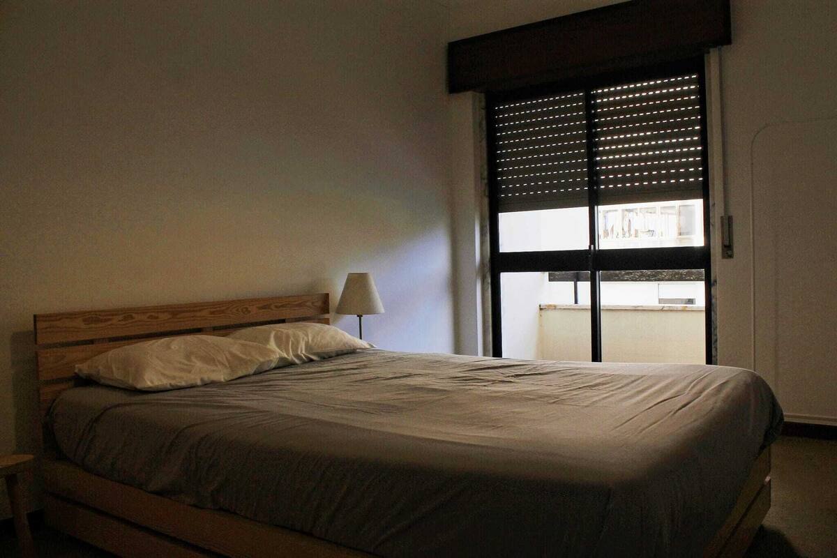 Quarto com cabeceira de madeira, uma cama de solteiro, mesa de cabeceira, luminária e janela com persianas.