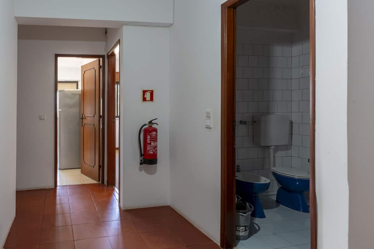 Um corredor com um extintor fixado na parede, uma porta que dá para um banheiro com vaso sanitário azul e pia branca.