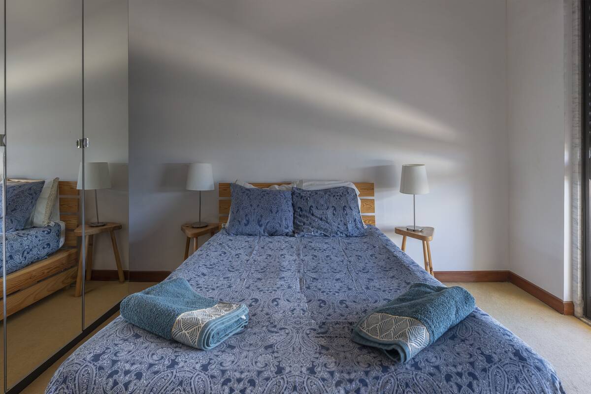 Quarto com uma cama grande, duas mesas de cabeceira com abajures, um espelho e uma cabeceira de madeira.