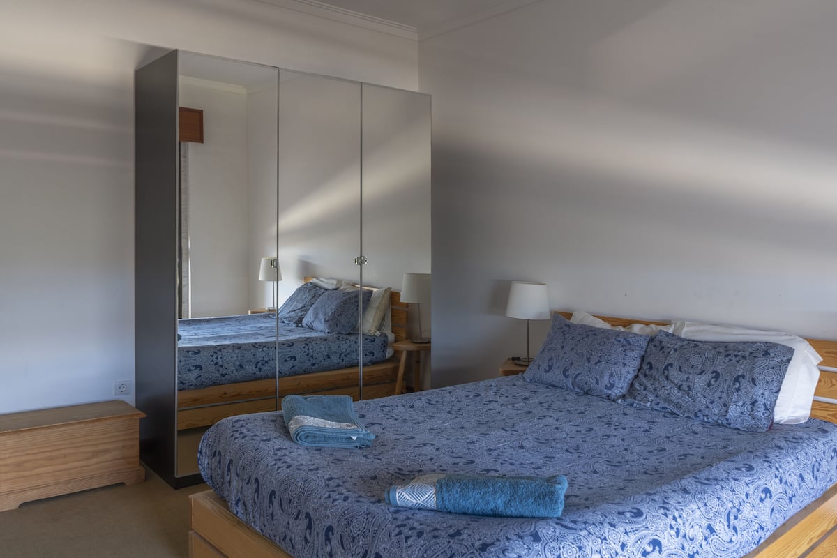 Quarto com um espelho grande, uma cama com roupa de cama estampada, um banco de madeira e uma mesa de cabeceira com abajur.