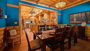 Interior de una cabaña de madera con comedor, cocina y un ventanal con vista al bosque.