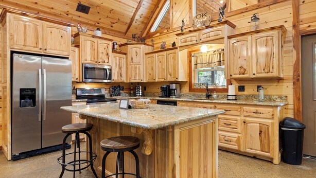A spacious kitchen with wooden cabinets, a stainless steel refrigerator, a microwave, and a coffee maker. There are two bar stools and a large island with a granite countertop.