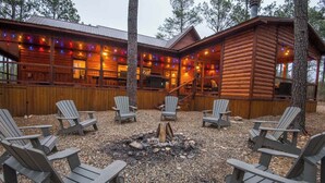 Una cabaña de troncos con un brasero rodeado de sillas Adirondack, una terraza de madera y luces colgantes.