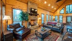 A spacious living room with a stone fireplace, leather furniture, and large windows offering views of the outdoors. - Fall in love with Legends Of The Fall cabin. Play Games & Soak in Large Hot Tub! (Broken Bow)