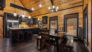 Interior rústico de una cabaña con cocina, comedor y amplias ventanas que ofrecen vistas al exterior.