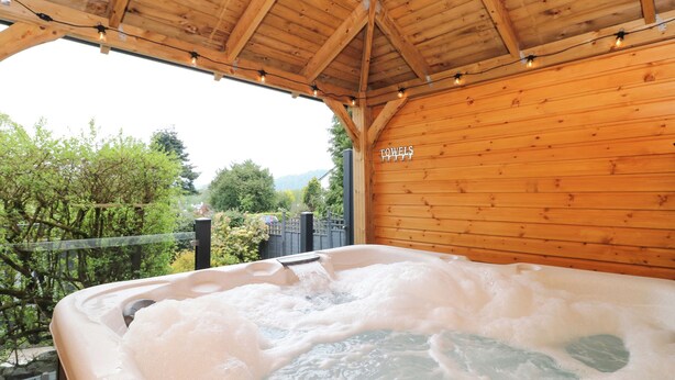 A hot tub under a wooden roof with a view of trees and a fence. - Briars Lea (Windermere)