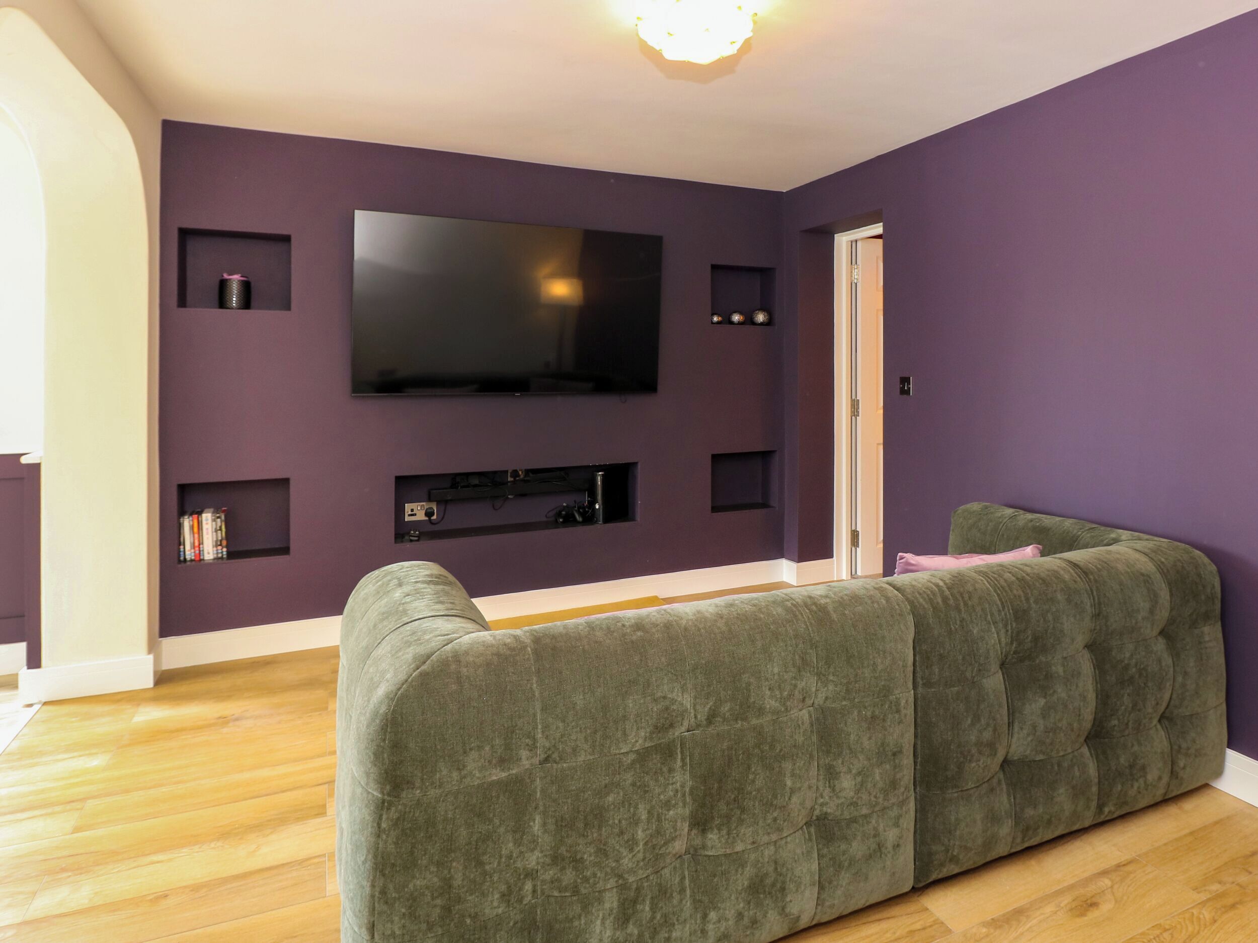 A living room with a large flat-screen TV mounted on a purple wall, a green sofa, and a wooden floor.