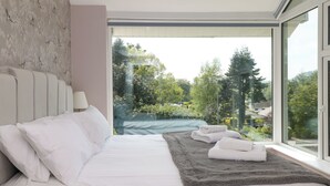 A modern bedroom with a large window, a bed with white linens, a grey rug, and a view of a garden. - Briars Lea (Windermere)