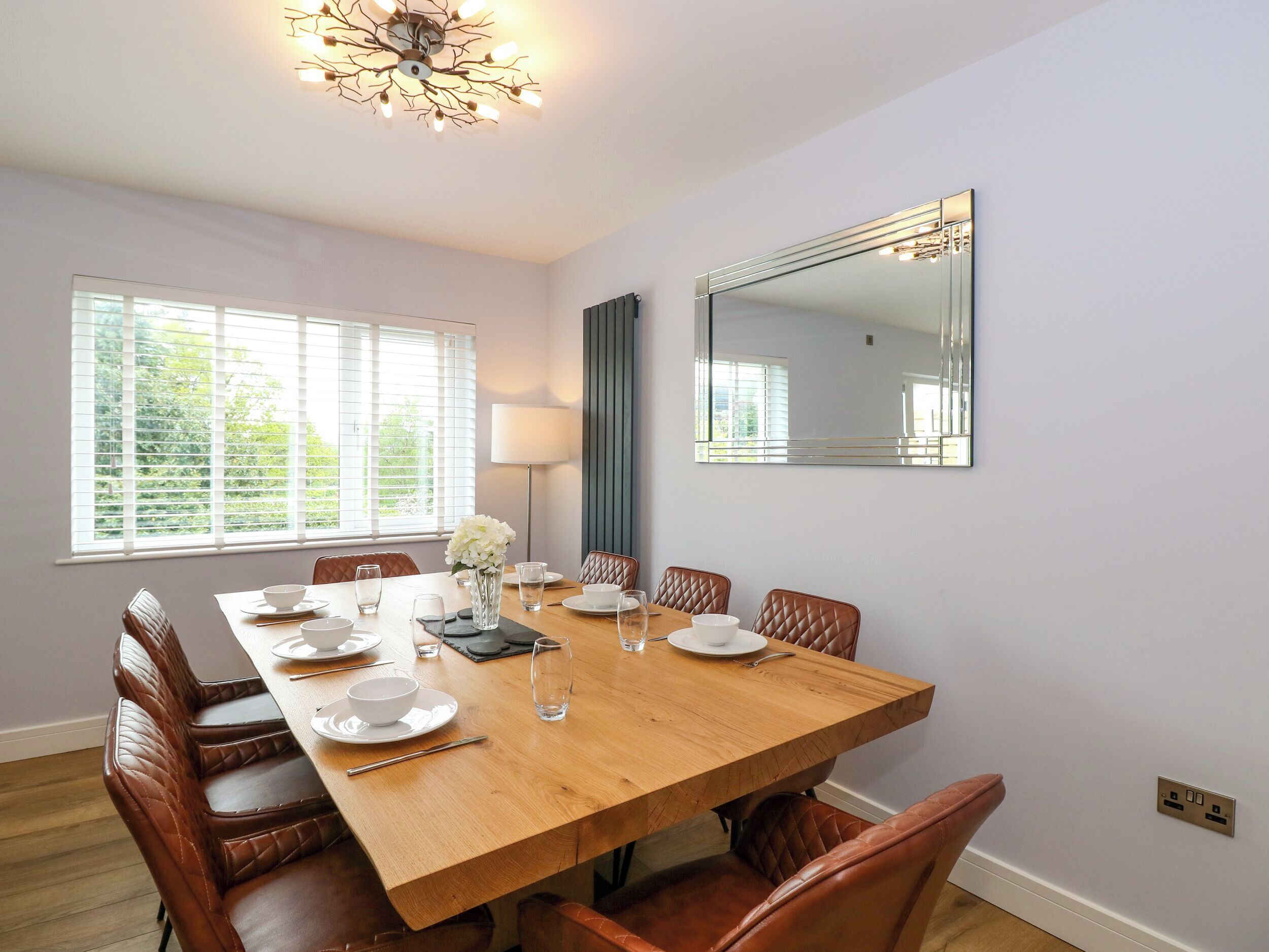 Une salle à manger avec une table en bois dressée pour six personnes, entourée de chaises en cuir. On y trouve une grande fenêtre avec des stores, un lampadaire et un miroir fixé au mur.