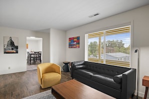 A modern living room with a black leather sofa, a yellow armchair, and a wooden coffee table. There is a dining area with a table and chairs, and a colorful abstract painting on the wall. - Flagstaff Getaway Triplex 6 Bedrooms, 4.5 Baths, Fenced Yards, AC, 3-Car Garage (Flagstaff)