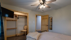 Un dormitorio con cama, ventilador de techo, armario de madera, silla y televisor montado en la pared.